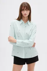 Blusa de corte holgado en color verde agua claro con rayas verticales finas y pequeños bordados florales dispersos. Presenta cuello camisero, cierre frontal con botones y mangas largas con puños abotonados.