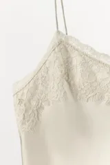 Top lencero color beige claro, confeccionado en mezcla de seda, con escote en pico y tirantes finos. Presenta una aplicación de encaje a tono en el escote.