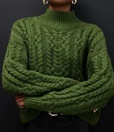 Suéter de punto color verde oliva con diseño de trenzas, cuello alto y mangas largas. Presenta un corte corto con terminaciones acanaladas en puños, cuello y ruedo.
