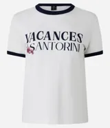 Blusa baby look de algodón color blanco, con cuello redondo y mangas cortas con ribetes azul marino. Presenta un estampado frontal con el texto "VACANCES SANTORINI" en azul marino y detalles florales en rosado.