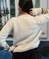 Sweater de lana merino tejido a mano, color marfil, con diseño jacquard geométrico en tonos marrón, violeta y gris en el pecho y las mangas. Cuenta con cuello redondo y mangas largas.