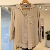 Camisa beige de manga larga, con cuello solapa, cierre frontal de botones y un bolsillo aplicado en el pecho.