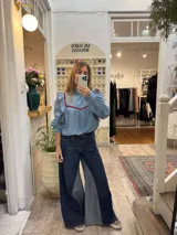 Blusa de jean fino con cuello alto, mangas largas con volumen y volados decorativos en los hombros. Presenta detalles de ribetes en terciopelo azul oscuro en el frente y los volados.