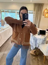 Cardigan de punto color beige con escote en V, cierre frontal con botones, bolsillos delanteros aplicados y detalle de flecos en el ruedo.