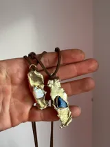 Collar artesanal con dije de base metálica irregular y piedra central roja. Presenta una estructura de tiento de gamuza marrón con detalles de tubos metálicos dorados.
