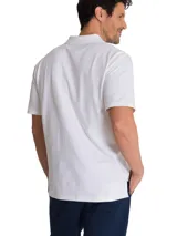 Remera tipo polo de manga corta color blanco, con cuello de punto acanalado y tapeta con botones. Presenta el logo de la marca CAT bordado en el pecho y un detalle de triángulo amarillo en la manga.