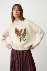 Sweater de punto color crudo, con cuello redondo y diseño de bordado floral en el frente. Presenta mangas largas con puños acanalados y un corte holgado.