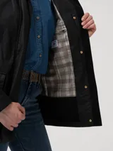 Chaqueta de algodón orgánico encerado, resistente al agua y al viento, con diseño de corte recto. Presenta cuello alto, gorro removible con forro a cuadros, cierre frontal metálico oculto por solapa con botones a presión y dos bolsillos frontales con solapa. Incluye detalles en cotelé y forro interior funcional.