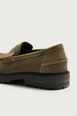 Mocasines de vestir color marrón oscuro, confeccionados en piel con acabado en gamuja serraje. Presentan detalle de antifaz en el empeine con costuras pespunteadas, punta redonda y suela track de goma robusta.