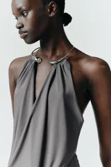 Vestido largo de color negro con cuello halter y detalle de aro metálico en el escote. Presenta un diseño drapeado en la parte frontal y escote en pico pronunciado.