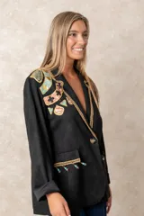 Blazer negro de corte recto con solapas decoradas con ribete dorado trenzado. Presenta bordados artesanales coloridos en el hombro derecho y detalles de flecos con cuentas turquesas en los bolsillos frontales. Cuenta con cierre de un solo botón.