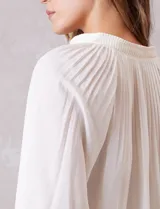 Blusa de gasa con plisado soleil, cuello a la base fruncido con escote en V regulable mediante lazos, y mangas largas abullonadas con puños sastreros.