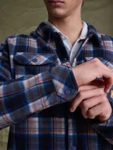 Sobrecamisa de tejido polar con estampado abstracto en tonos rojizos y azul marino. Presenta cuello clásico, cierre frontal con botones a presión, dos bolsillos frontales con solapa en el pecho y un parche decorativo.