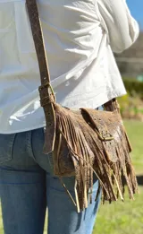 Mini bandolera de cuero con diseño de flecos largos y hebilla frontal decorativa. Presenta un acabado texturizado con efecto craquelado en tonos blanco y marrón, y correa ajustable.