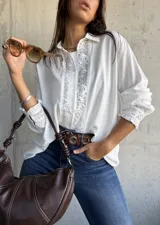 Camisa blanca de corte clásico con detalles de encaje floral vertical en la parte frontal y en los puños. Presenta cuello camisero, cierre frontal con botones y mangas largas con terminación elástica.