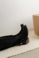 Bota estilo tejana de caña alta, confeccionada en cuero marrón con acabado texturizado. Presenta punta cuadrada, taco bajo y una correa con hebilla metálica ajustable y estribo desmontable en la zona del tobillo.