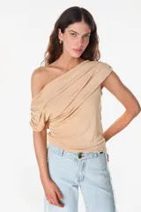 Blusa asimétrica color beige de poliéster con spandex, con diseño drapeado envolvente que deja un hombro al descubierto y manga corta caída del otro lado.