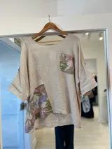 Blusa beige de lino con diseño asimétrico, mangas cortas y detalles de tela estampada con motivos florales en el bolsillo y en la parte inferior.