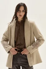 Blazer de paño color beige, con solapas clásicas y bordados decorativos en tono más oscuro en la parte inferior de las mangas.