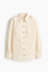 Camisa de estilo cargo color beige, confeccionada en mezcla de lino y algodón. Presenta cuello clásico, cierre frontal con botones, bolsillos superiores con solapa, charreteras en los hombros y mangas largas con puños abotonados.