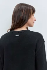 Cardigan negro de punto con acabado brillante, cuello redondo, cierre frontal de botones y detalle de cordones laterales entrelazados. Presenta mangas tres cuartos con diseño acampanado.