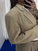 Conjunto de dos piezas color beige compuesto por un blazer corto con apliques metálicos tipo alfileres y un short con detalle de puntilla blanca en el ruedo.