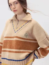 Pullover de punto con textura aterciopelada, diseño oversize, cuello polo con cierre frontal parcial y detalles de pespuntes en contraste. Presenta rayas horizontales en tonos tierra y un parche bordado con la letra M en el pecho.