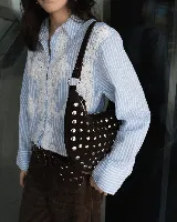Bolso de hombro tipo hobo, confeccionado en gamuza color marrón oscuro, decorado con tachas metálicas plateadas en toda su superficie frontal. Cuenta con un asa corta fija y cierre superior.