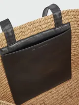Tote bag de rafia natural con diseño tejido, asas largas de cuero negro y bordado frontal con el logo de la marca en hilo negro.
