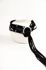 Cinturón negro de cuero con hebilla metálica plateada en forma de aro. El cinturón se anuda y tiene una tira de gamuza negra con flecos colgantes.