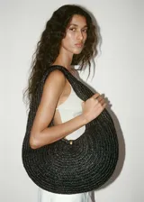 Bolso tipo hobo de diseño ovalado, confeccionado en tejido de fibra de papel trenzado en color negro. Presenta un asa integrada al cuerpo del bolso y un cierre central mediante un botón metálico dorado.