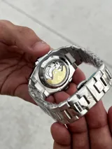 Reloj de pulsera con diseño inspirado en el modelo Nautilus, con caja y malla de acero inoxidable. Presenta una esfera azul con textura estriada horizontal, índices aplicados luminiscentes y ventana de fecha a las tres. Incluye cierre desplegable tipo mariposa.