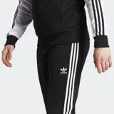 Pantalón deportivo Adidas Adicolor Classics SST negro con tres rayas blancas a los costados, corte ajustado, puños acanalados, logo del trifolio bordado en la pierna izquierda, bolsillos laterales con cierre y cintura elástica con cordón de ajuste.