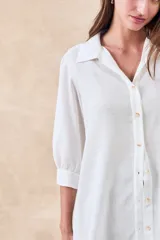 Camisa de corte holgado y fluido, color blanco, con cuello clásico, cierre frontal de botones y mangas tres cuartos con puños rectos.