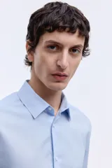 Camisa de tela azul claro con tratamiento de planchado fácil, cuello inglés, tapeta clásica y canesú en la espalda. Mangas largas con tapeta y botón, y cierre ajustable de botón en los puños. Bajo levemente redondeado. Corte ajustado que realza el contorno del cuerpo.