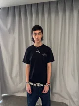 Remera de algodón color negro con estampado serigráfico en tono gris oscuro en la parte posterior. Presenta cuello reforzado, costuras reforzadas y una pequeña etiqueta decorativa en la manga.