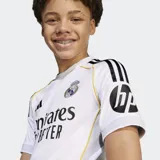 Camiseta de fútbol Adidas del Real Madrid, primera equipación temporada 25/26. Diseño en color blanco con detalles en negro y líneas amarillas sutiles, cuello redondo y mangas cortas. Incluye el escudo del club bordado y el logo de la marca.