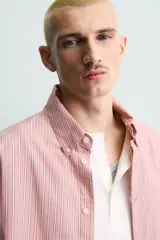 Camisa de corte relajado confeccionada en tejido de algodón, con diseño de rayas verticales en tonos rosados y blancos. Presenta cuello abotonado, cierre frontal de botones, manga larga con puños abotonados y bajo asimétrico.
