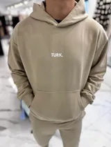 Conjunto de jogging beige que incluye un hoodie con capucha, bolsillo frontal tipo canguro y pequeño texto estampado en el pecho, acompañado de un pantalón tipo jogger con elástico en los tobillos.