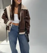 Campera tipo aviador de cuero sintético color marrón camel, con interior y bordes de corderito sintético color crema. Presenta cierre frontal, detalles de correas con hebillas en el cuello y en el ruedo, y ribetes negros en las costuras.