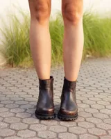 Bota de caña alta confeccionada en cuero color marrón caoba con acabado desgastado. Presenta una suela track robusta, correas con hebillas en la parte superior de la caña y un arnés decorativo con herrajes metálicos en el tobillo.