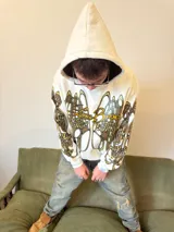 Hoodie oversize de color blanco con estampado artístico en tonos marrones y dorados en el pecho y las mangas. Presenta un forro interior de capucha en tejido marrón texturizado y costuras reforzadas.