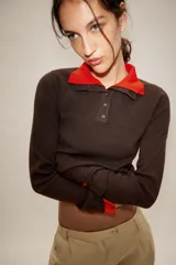 Jersey de punto con efecto doble, cuello subido y manga larga. Presenta un diseño en color marrón con detalles en rojo vibrante en el cuello, puños y ruedo. Cuenta con cierre frontal parcial mediante botones.