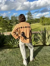 Buzo tejido de punto color beige, con diseño estampado de un caballo y rombos en la parte superior trasera. Presenta cuello redondo, mangas largas y corte holgado.
