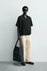 Camisa de corte relajado en color negro, confeccionada en tejido de algodón. Presenta cuello tipo bowling, manga corta y cierre frontal con botones. Cuenta con bordados lineales de hojas en la parte delantera.