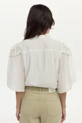 Blusa blanca con cuello mao, detalles de mini volados y bordados en el pecho y las mangas, con cierre frontal de botones y mangas largas abullonadas.