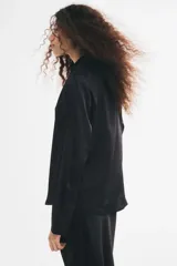 Blusa negra de tela brillante, con cuello clásico, escote en V con botones frontales, mangas largas con puños sencillos y bajo redondeado.