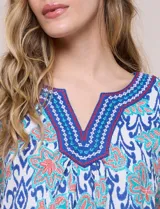 Blusa de manga corta con escote en V y pechera bordada. Presenta un estampado multicolor sobre base blanca, con pliegues frontales y aberturas laterales en el ruedo.