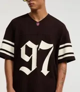 Remera deportiva de algodón color marrón con cuello en V y mangas cortas. Diseño overboxy con estampado de fútbol americano, lettering y rayas contrastantes en blanco.