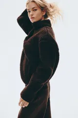 Buzo de sherpa con textura de peluche, cuello elevado y medio cierre frontal. Presenta hombros caídos, mangas largas y bolsillos laterales insertados.